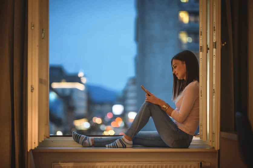 Ung dame som sitter i vinduskarmen mens hun ser på mobilen sin